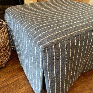 May include: A square ottoman upholstered in blue fabric with vertical white stripes. The fabric has a textured appearance, and the ottoman sits on a wooden floor. The design is simple and classic, suitable for various interior styles.