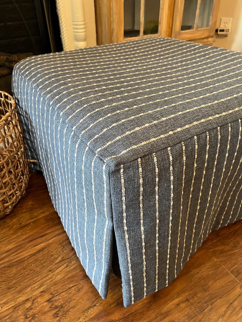 May include: A square ottoman upholstered in blue fabric with vertical white stripes. The fabric has a textured appearance, and the ottoman sits on a wooden floor. The design is simple and classic, suitable for various interior styles.