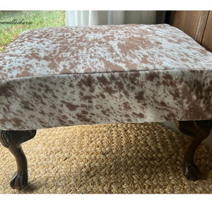May include: A brown and white cowhide ottoman with four wooden legs. The ottoman is sitting on a woven rug.