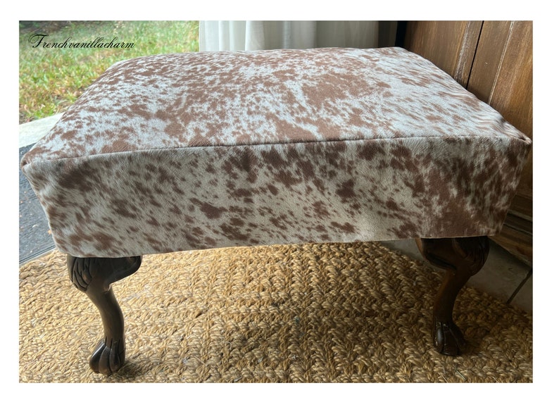 May include: A brown and white cowhide ottoman with four wooden legs. The ottoman is sitting on a woven rug.