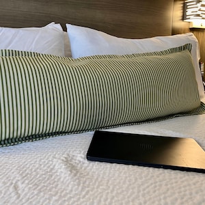 May include: A long rectangular bolster pillow featuring a green and white pinstripe pattern, positioned horizontally across a white textured bedspread. A closed black laptop computer rests on the bedspread in front of the bolster pillow.