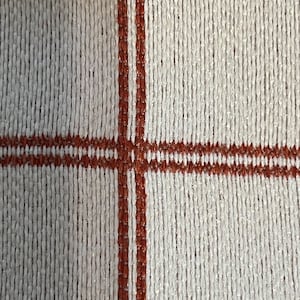 May include: A close-up of a white woven fabric with two brown stripes running vertically and horizontally across the fabric.