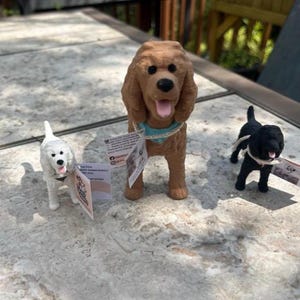 May include: Three dog figurines including a small white dog, a larger brown dog with a light blue collar, and a small black dog. Each figurine features a small white tag attached to its neck area, displaying printed text and social media icons.