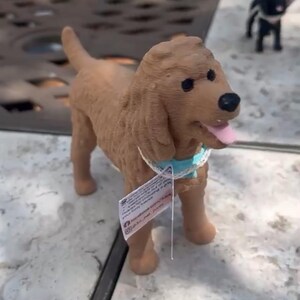 May include: A brown dog figurine with a textured coat, black eyes, and a black nose. The dog has a pink tongue visible and wears a light blue collar with a white lace trim. A small white rectangular tag with printed text hangs from the collar.