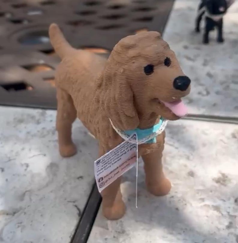 May include: A brown dog figurine with a textured coat, black eyes, and a black nose. The dog has a pink tongue visible and wears a light blue collar with a white lace trim. A small white rectangular tag with printed text hangs from the collar.