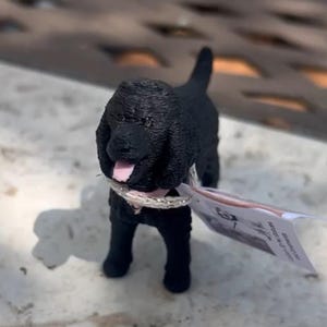 May include: A small black dog figurine with a textured coat, floppy ears, and an open mouth showing a pink tongue. The dog wears a thin, metallic silver-colored collar around its neck. A small white paper tag is attached to the collar.