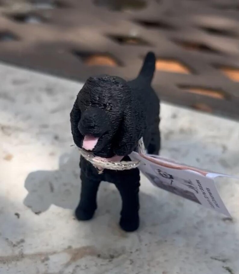 May include: A small black dog figurine with a textured coat, floppy ears, and an open mouth showing a pink tongue. The dog wears a thin, metallic silver-colored collar around its neck. A small white paper tag is attached to the collar.
