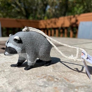 Pu&ograve; includere: Una piccola statuetta tridimensionale di procione con corpo grigio testurizzato, maschera nera sugli occhi e zampe nere. Un occhiello in metallo argentato &egrave; fissato sulla parte superiore, collegato a un cordino beige con un piccolo cartellino.
