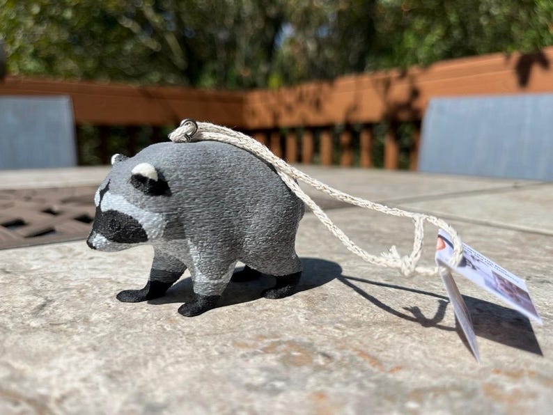 Pu&ograve; includere: Una piccola statuetta tridimensionale di procione con corpo grigio testurizzato, maschera nera sugli occhi e zampe nere. Un occhiello in metallo argentato &egrave; fissato sulla parte superiore, collegato a un cordino beige con un piccolo cartellino.