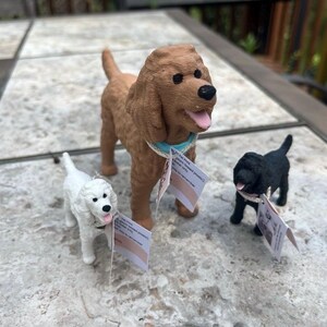 May include: Three dog figurines in brown, white, and black. The brown dog wears a light blue collar, the white dog has a dark collar, and the black dog has a light collar. Each figurine features a small white tag with printed text attached.