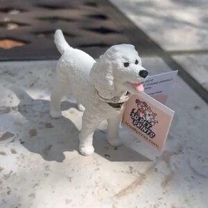 May include: A white dog figurine with a textured coat, a black nose, and a pink tongue. The dog wears a silver-colored collar and has a small white tag attached that features a graphic of two dogs and the text 3D PET PRINTS 3dpetprints.com.