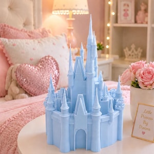 Puede incluir: Una figura de castillo azul claro con múltiples torres se encuentra sobre una mesa de noche blanca. El dormitorio tiene una decoración rosa, incluyendo ropa de cama y una lámpara. Un letrero enmarcado dice "Little Princess Big Dreams."