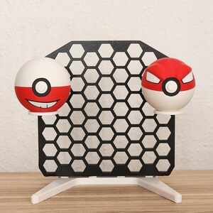 Può includere: Un espositore con due Poké Ball rosse e bianche. Il supporto ha un design a nido d'ape nero e una base bianca. Una Poké Ball ha una faccia sorridente, l'altra una faccia arrabbiata.