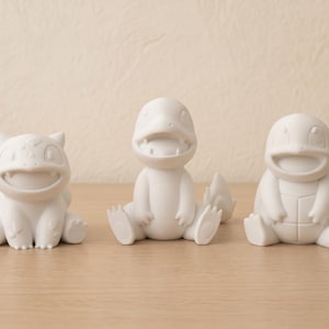 Puede incluir: Tres figuras de personajes en blanco mate dispuestas en fila. La figura de la izquierda tiene una espalda bulbosa, la del centro tiene una cola larga y la boca abierta, y la de la derecha presenta una espalda con forma de caparazón.