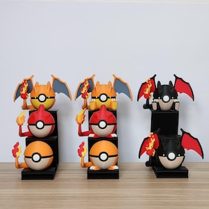 Op de afbeelding: Drie sets Pokémon-figuren. Elke set bevat een Charizard-figuur met oranje, rode en zwarte kleurenschema's, die een vlam vasthoudt. De figuren staan op zwarte display stands, met een Poké Ball-ontwerp.