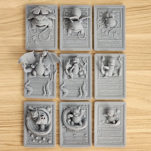 Könnte beinhalten: Neun graue, 3D-gedruckte Pokémon-Sammelkarten mit verschiedenen Charakteren wie Charizard, Blastoise und Squirtle. Jede Karte hat ein erhabenes Reliefdesign des Pokémon und seines Namens sowie weitere Details. Die Karten sind auf einer hellen Holzmaserung angeordnet.