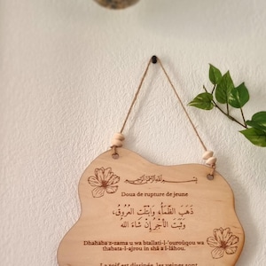 Puede incluir: Un letrero de madera con una oración islámica para romper el ayuno durante el Ramadán. El letrero está decorado con diseños florales y tiene una cuerda para colgar. El texto en el letrero dice: "Dhahaba z-zama u wa btallati-l-'ouroûqou wa thabata-l-ajrou in shâ a l-lâhou. La soif est dissipée, les veines sont abreuvées et la récompense restera avec la volonté d'Allah."