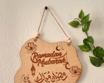 May include: A wooden sign with a brown background and a white inscription that reads "Ramadan Mubarak" in English and Arabic. The sign is decorated with a crescent moon, stars, and flowers.