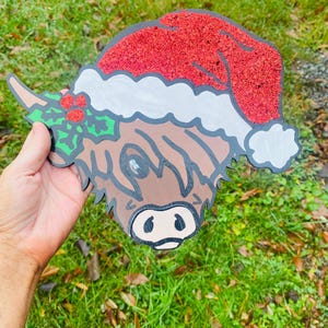 Puede incluir: Una cabeza de vaca de las Tierras Altas de madera con un gorro de Papá Noel con purpurina roja y ribete blanco. La vaca tiene pelaje marrón, un contorno negro y una ramita de acebo con bayas rojas. Una decoración navideña festiva.