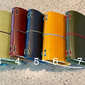 May include: Eight leather journals arranged in a row, numbered one through eight. Each journal features a distinct color, including brown, green, light green, red, navy, yellow, olive, and burgundy, secured with a thin elastic band and metal eyelets.