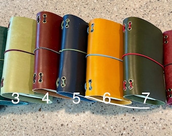 May include: Eight leather journals arranged in a row, numbered one through eight. Each journal features a distinct color, including brown, green, light green, red, navy, yellow, olive, and burgundy, secured with a thin elastic band and metal eyelets.