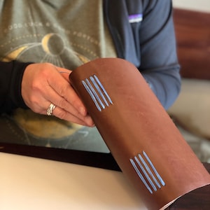 May include: A brown leather journal with blue stitching. The journal is being held by a person's hand.