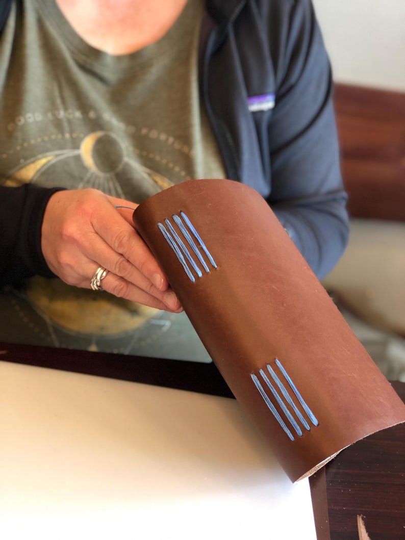 May include: A brown leather journal with blue stitching. The journal is being held by a person's hand.