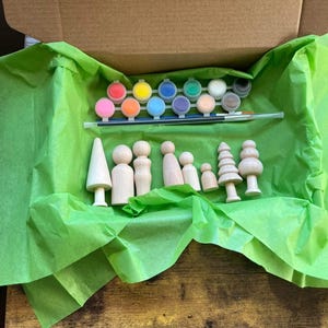 May include: A green gift box filled with wooden figurines and paint pots. The figurines include a family of people and trees. The paint pots are in a variety of colors, including red, blue, green, yellow, and orange.