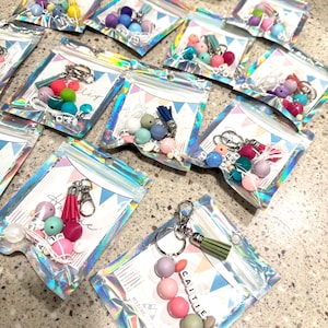 May include: A collection of colorful beaded keychains in clear, iridescent pouches. Each keychain features silicone beads in various pastel colors, a tassel, and a metal clasp. Some keychains have names spelled out with letter beads.