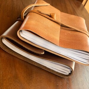 May include: A brown leather-bound journal with a tan leather cover and a strap closure. The journal is open to reveal blank pages.