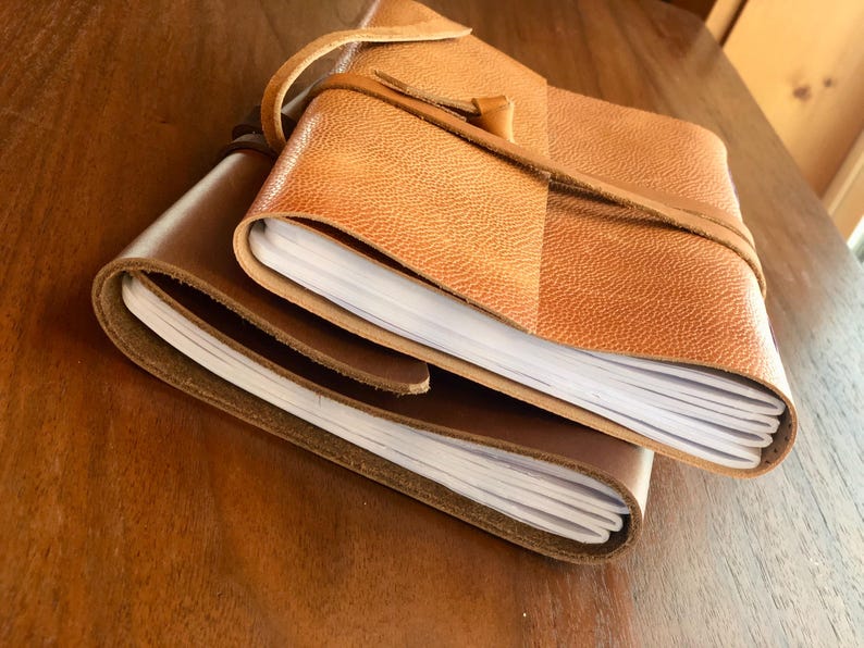 May include: A brown leather-bound journal with a tan leather cover and a strap closure. The journal is open to reveal blank pages.