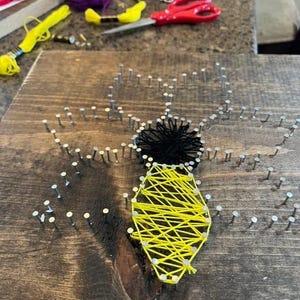 May include: A handmade string art piece featuring a bee design. The bee's body is crafted with yellow string, and the head is black. The artwork is created on a stained wooden board with small nails outlining the bee's shape. Craft supplies are visible in the background.