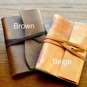 May include: Two leather-bound journals, one brown and one beige, with leather straps and ties. The brown journal has the word "Brown" written on it, and the beige journal has the word "Beige" written on it.