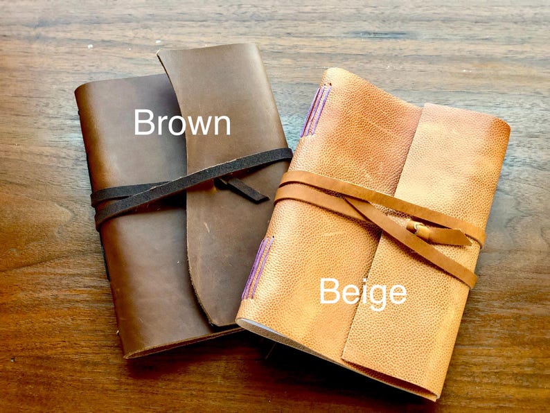 May include: Two leather-bound journals, one brown and one beige, with leather straps and ties. The brown journal has the word "Brown" written on it, and the beige journal has the word "Beige" written on it.