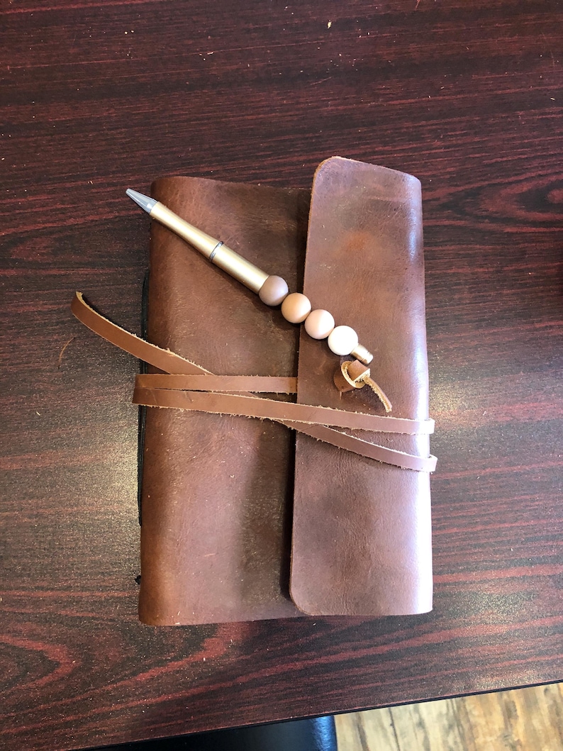 May include: A brown leather journal with a pen and a beaded tassel. The journal is closed and tied with leather straps.