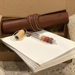 Può includere: Un diario in pelle marrone con una penna e un rotolo di filo all'interno di una scatola di cartone. La penna ha un fusto argentato e un set di perline colorate.