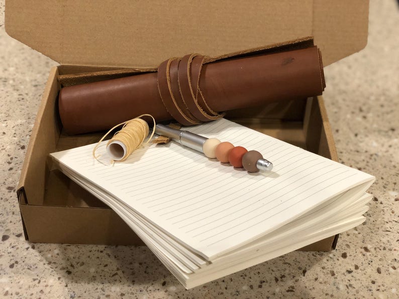 May include: A brown leather journal with a pen and a spool of thread inside a cardboard box. The pen has a silver barrel and a set of colorful beads.