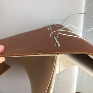 May include: A close-up of a leather-bound book being sewn together with a needle and green thread. The book is open and the pages are visible.