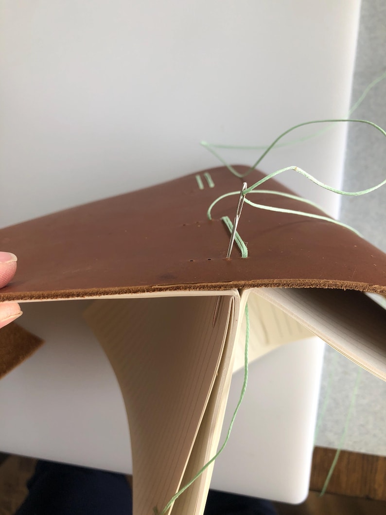 May include: A close-up of a leather-bound book being sewn together with a needle and green thread. The book is open and the pages are visible.