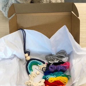 May include: A rainbow-colored yarn craft kit in a cardboard box. The kit includes yarn in various colors, wooden beads, and a needle.