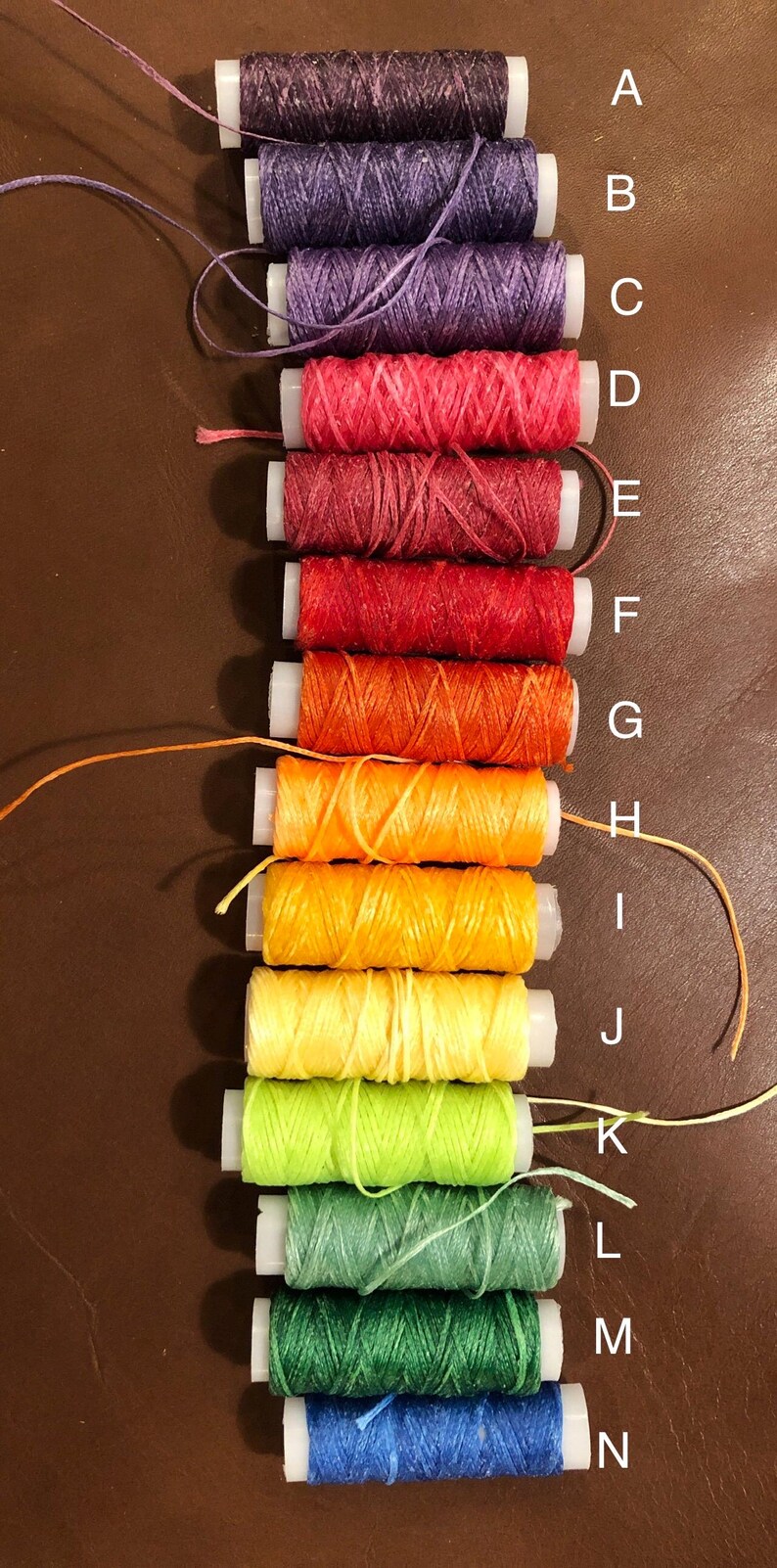 May include: A set of 14 spools of thread in various colors, including purple, red, orange, yellow, green, and blue. The spools are labeled with letters from A to N.