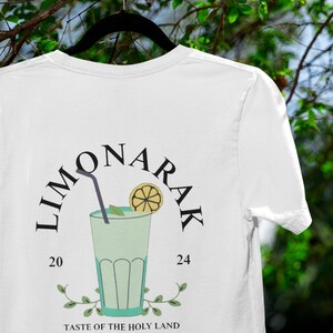 May include: White t-shirt with a black graphic of a glass of lemonade with a lemon slice and a straw. The graphic says "LIMONARAK 20 24" and "TASTE OF THE HOLY LAND".