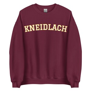 Könnte beinhalten: Burgunderfarbener Pullover mit dem Text "KNEIDLACH" in Gelb.