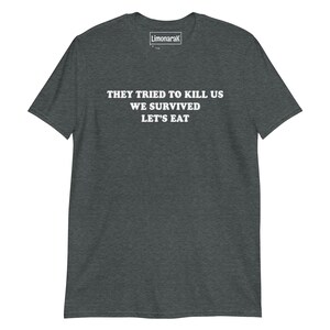 May include: Dark gray t-shirt with white text that reads "THEY TRIED TO KILL US WE SURVIVED LET'S EAT". The shirt has a crew neck and short sleeves. The brand name "Limonarak" is printed on the upper right.