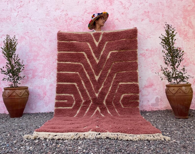 May include: A pink and cream patterned rug with a geometric design. The rug has a fringe border and is laid out on a pink and gray surface.