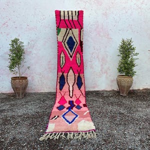 May include: A vibrant, rectangular rug with a pink and white geometric design. The rug features diamond and oval shapes in blue, pink, and green. The rug has a fringe at the bottom and is displayed outdoors.