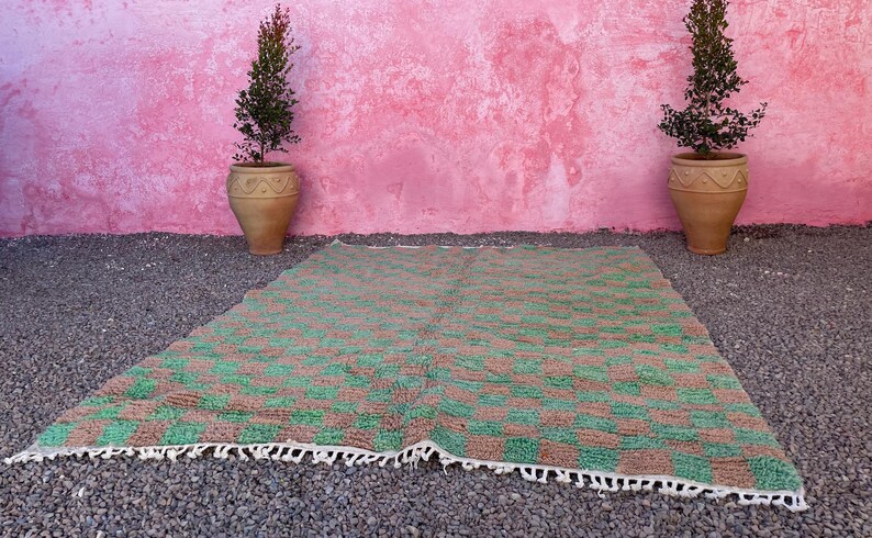 May include: A green and brown checkered rug with a white fringe border. The rug is laid out on a gravel surface in front of a pink wall with two potted green plants.
