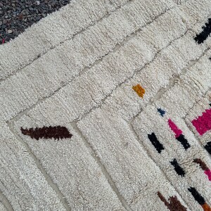 May include: Cream-colored, textured rug with a geometric design. The rug features rectangular and diagonal patterns with accents of brown, black, pink, yellow, and blue. The rug's design is visible in this close-up shot.