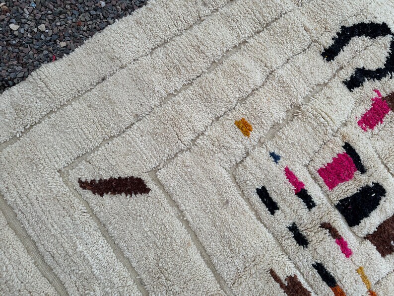 May include: Cream-colored, textured rug with a geometric design. The rug features rectangular and diagonal patterns with accents of brown, black, pink, yellow, and blue. The rug's design is visible in this close-up shot.