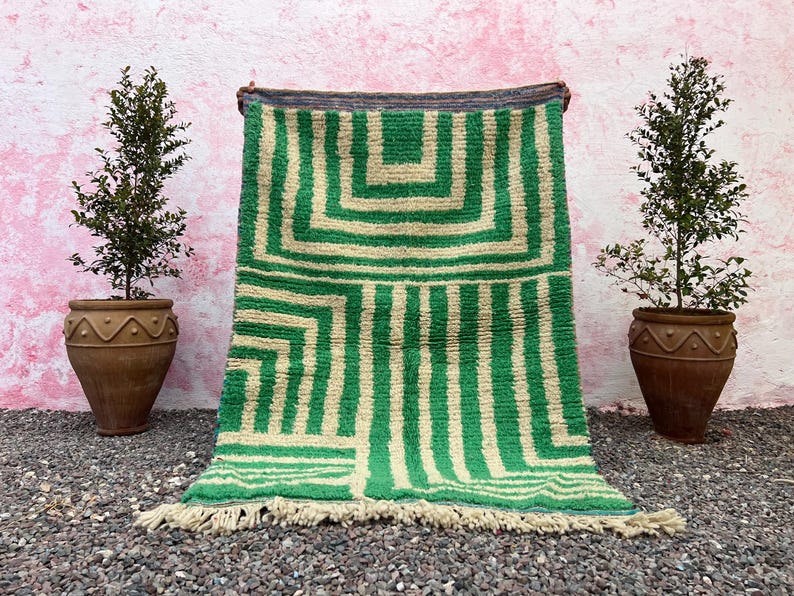 Pode incluir: Um tapete com padr&atilde;o geom&eacute;trico verde e creme com borda com franjas. O tapete &eacute; exibido contra uma parede rosa, ladeado por duas plantas em vasos de terracota marrom. O design do tapete apresenta quadrados e listras verticais.